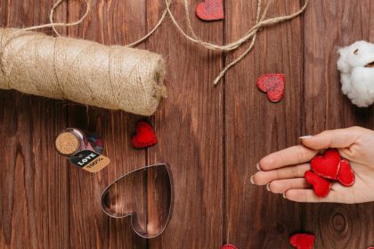 A wooden table surface. A hand hold out three homemade red hearts. A roll of string and hearts, including a cookie cutter, lie on the table
